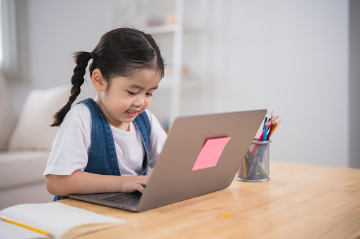 Asian baby girl smiling use laptop or writing drawing color on note book study online on wood table desk in living room at home. Education learning online from home concept.