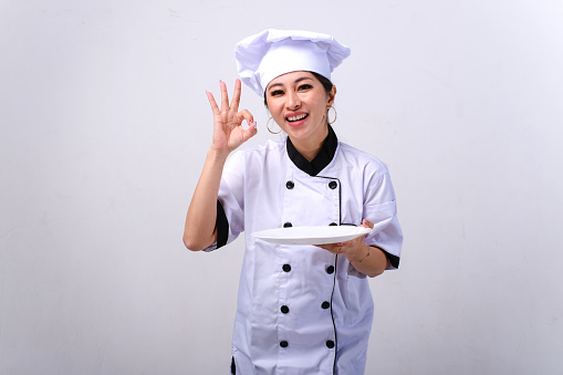Smiling female Asian chef holding empty plate and showing OK hand sign gesture isolated on white background