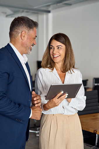 Vertical portrait of mature Latin businessman and European businesswoman discussing project on tablet in office. Two diverse partners colleagues of confident professional businesspeople work together.