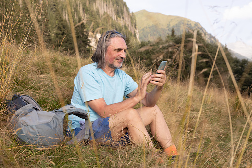A gray-haired long-haired man with a backpack sits in the tall grass against the backdrop of mountains and communicates on a mobile phone. High quality photo