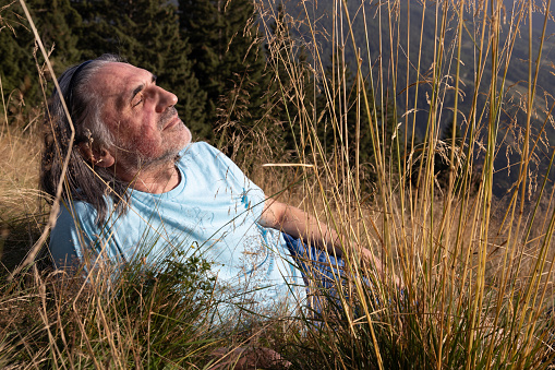 Adult gray-haired man with long hair resting on a field with tall grass in the evening sunlight. High quality photo