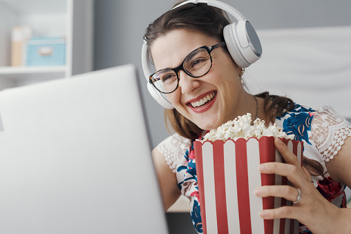 Happy woman sitting at her desk at home, she is watching online movies on the laptop and eating popcorn