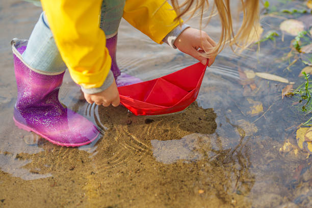 glückliches kindermädchen in gelbem regenmantel und papierboot am fluss im herbst auf natur. - puddle rain boot water stock-fotos und bilder