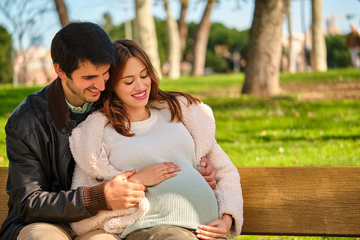 Happy pregnant couple embracing and touching her belly sitting on a bench in a park.