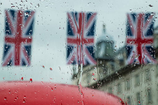 Three Union Jack flags are seen through the upper deck window of a London double decker bus on a rainy day.