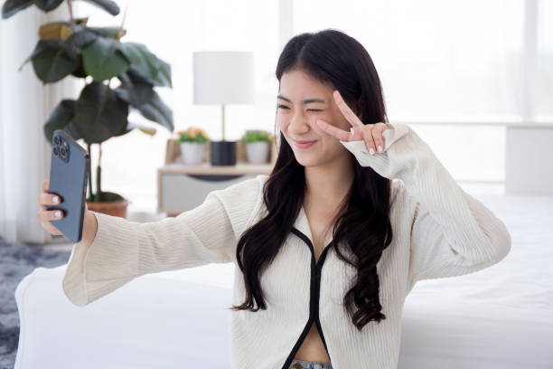 Beautiful young asian woman selfie with smartphone on social media in bedroom at home, girl posing and take a photo for relax, teenage leisure in room with happiness, lifestyles concept. stock photo