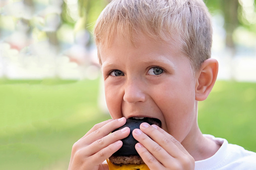 The child eats a black burger on the street. The boy bites off pieces of a cheeseburger. Fast food