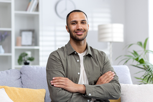 Portrait of a young Muslim man sitting on the couch at home and looking at the camera smiling, arms crossed.