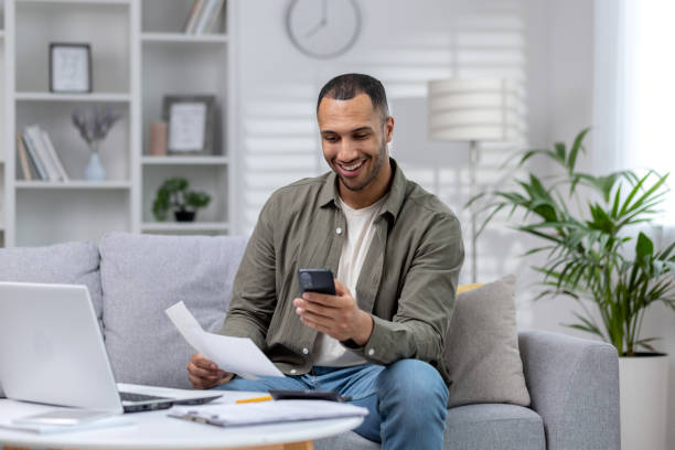 istock Young smiling African-American man sitting on sofa at home, holding phone and documents, working on laptop 1665396675