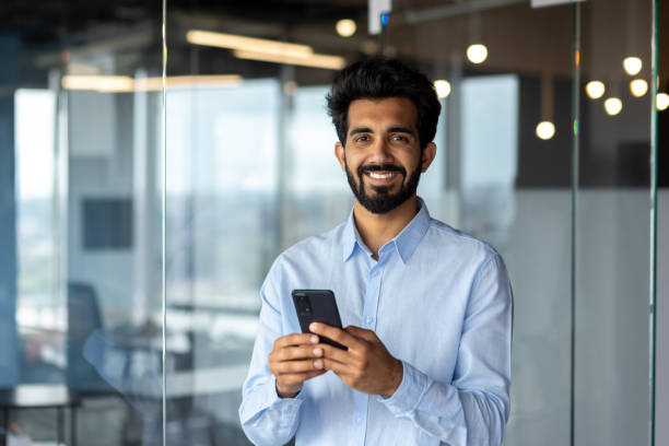 istock Portrait of Indian young male student standing inside campus, office, university and using phone. Smiling and looking at the camera 1665423679