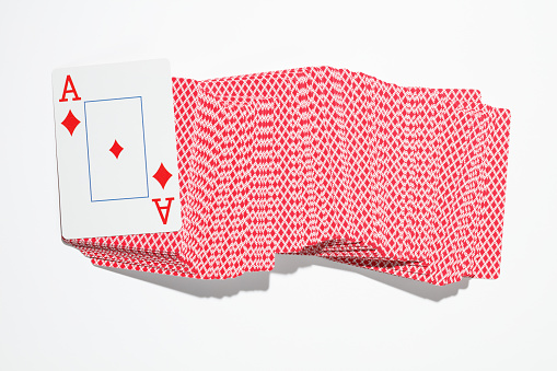 One shot glass rests on a black table surface in the upper right corner of this horizontal frame, with a hand of playing cards fanned out adjacent to it at left. \nThe card hand consists of the three of hearts atop the five of diamonds, atop the ten of spades, atop the jack of hearts, atop one card turned down.  Bright but filtered lighting enters the top of frame, casting a prism-like shadow from the shot glass onto the dark table top.  A moody, contrasty, old-style Wild West image.