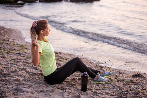 Young woman in sportswear is sitting on the seashore at sunrise. Girl is resting after workout, replenishes her water balance, drinks from a sports bottle, looks at the water. Morning outdoor fitness classes.