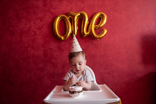 Little boy of one year old in party hat sits in childrens chair, eating birthday cake, Meringue, blows out candle. Kid on red isolated background, on textured wall foil gold balloons One. Home party