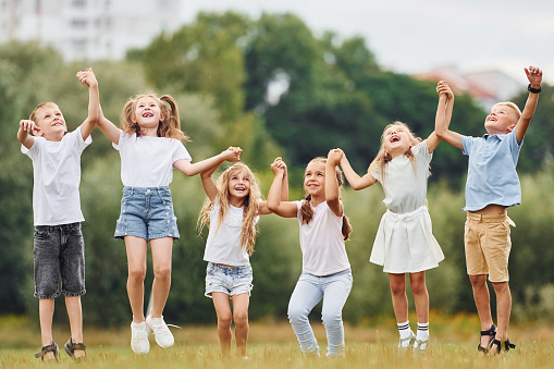 Holding each other by hands and jumping. Kids are having fun on the field at daytime together.