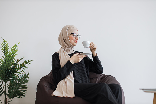 Portrait of beautiful woman in muslimah formal outfit drinking coffee while resting in soft pouf chair, copy space. Effective employee in glasses and heafscarf taking break during working day.