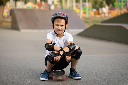 Cute kid boy child in a helmet sitting in a special area in skate park and holding skateboard. Summer sport activity concept. Happy childhood