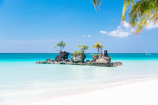 A tropical island - rock formation in the ocean. The island is small and has a few palm trees and rocks on it. The water is a beautiful shade of blue and is very clear. The sky is blue with a few clouds. The sand is white, and the beach is empty. The image is taken from the perspective of someone standing on the beach looking out at the island.