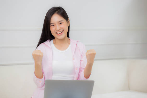 istock Happiness young asian woman sitting on sofa and working on laptop computer while successful and excitement at home, freelance female using notebook and achievement, business and lifestyles concept. 1665574404