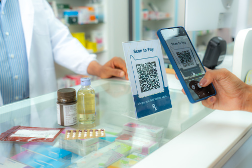 Unreconized patient's hand making a contactless payment at drug store by using smartphone to scan QR code from online banking after receiving medicine prescription from pharmacist man. Customer is making contactless payment.