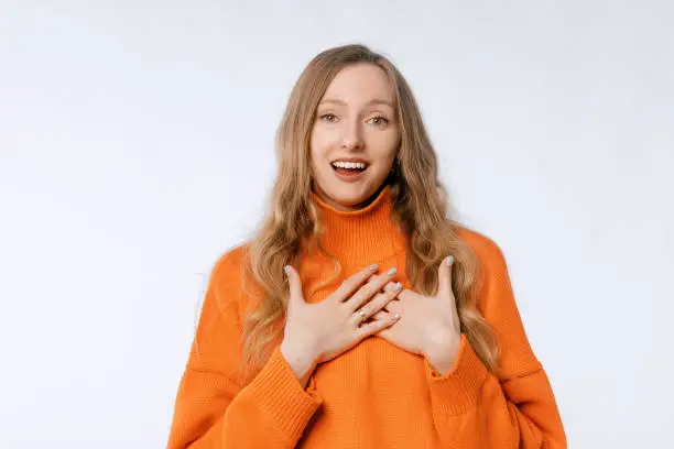 Smiling pretty woman, looking cute and flattered, standing trendy knitwear orange sweater over white background, holding hands on heart