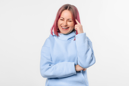 Pretty portrait of young woman thinking looking down and smiling having a finger at her head. Lovely female model with pink hair in a blue hoodie standing over white background