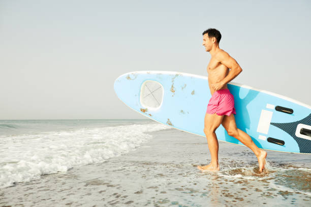 A man walking by the sea shore carrying a paddleboard.