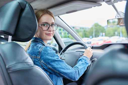 Teenage girl driver in glasses sitting behind wheel of car, smiling looking at camera, young female got license to drive car. Youth, auto school, lifestyle concept
