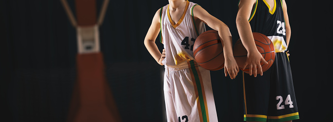 Two little boys holding basketball balls under their arms. Young athlete standing with a basketball under arm. School basketball players in basketball jersey shirts