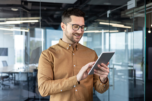 A young smiling male businessman in glasses is standing in the office and using a tablet. typing, chatting, searching for information.