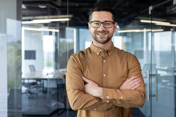 istock Portrait of a young male programmer, creative designer, developer engineer. He stands in the office with his arms crossed on his chest and looks at the camera with a smile 1665704151