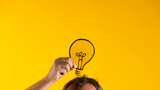 Hand holding light bulb on head against yellow background.