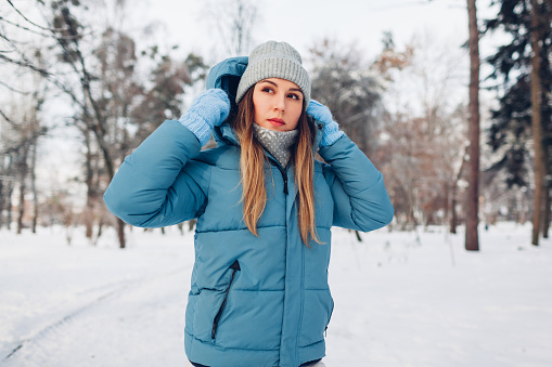 Portrait of young woman walking in snowy winter park wearing blue coat down jacket. Warm clothes for cold weather.