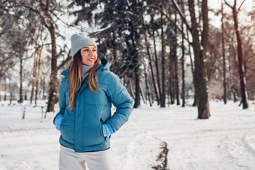 Young woman walking in snowy winter park wearing blue coat. Girl enjoys landscape wearing hat and mittens outdoors