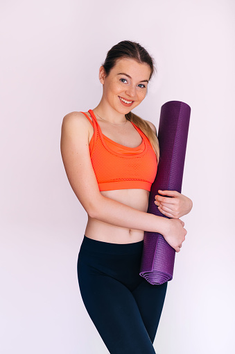Beautiful young woman in sportswear holds violet yoga mat, looking at camera and smiling, standing on white background