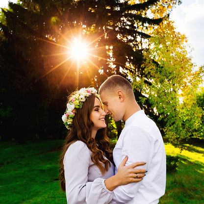 Husband hugs her beautiful wife with a wreath of flowers on her head against green trees and the sun