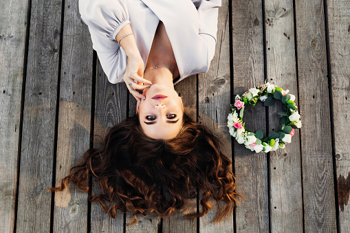 the woman lies on a wooden surface and looks at the camera lens. a flower wreath lies near girl