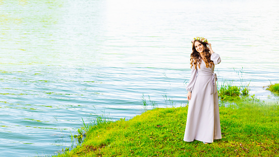 A girl in a long dress and wreath on her head walks on the shore of the lake