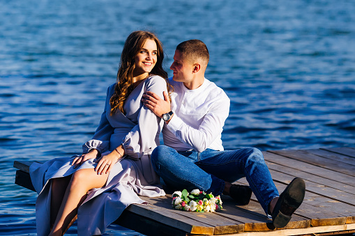 woman with long hair sitting on the pier and smiling and her husband sitting next to her and hugs her on a beautiful sunny day