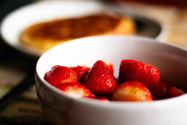 A bowl of ripe red strawberries on the table at breakfast with pancakes in the background. Morning meal detail, healthy fresh fruit.