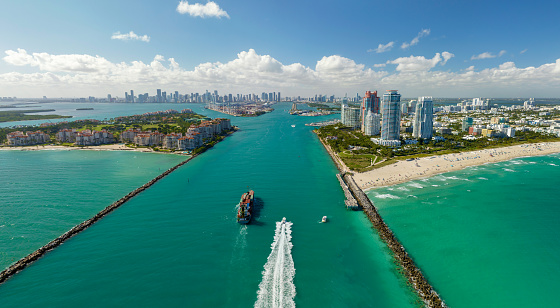Commercial container ship entering Miami port harbor through main channel near South Beach. Luxurious hotels and residential buildings on waterfront and high skyscraper towers of downtown in distance.