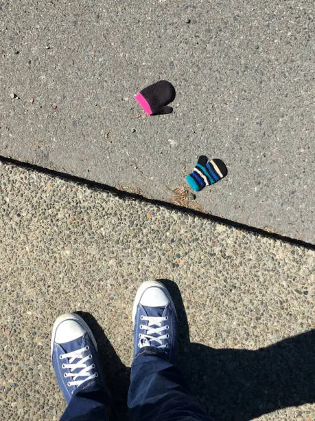 Person's point of view looking down on their shoes and two lost gloves while standing on a sidewalk.