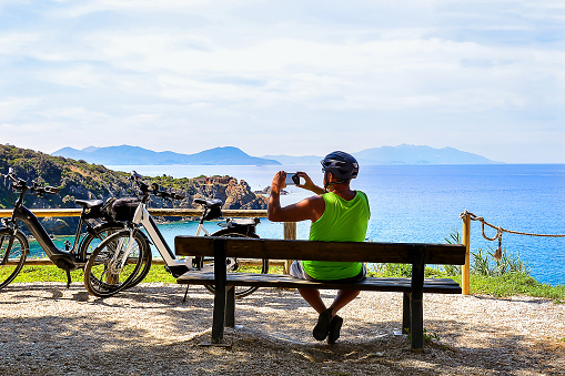 A 52-year-old man is resting in the shade on a wooden bench and taking pictures with a black phone against the backdrop of the Tyrrhenian sea and mountains in Italy, with two bikes standing nearby. Elba Island is visible opposite.