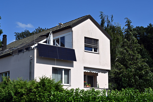 Urdorf, Switzerland 07 30 2022: Solar panels or photovoltaic plates installed on the balcony of family house observed in a Switzerland village. There are trees and bushes around the house.