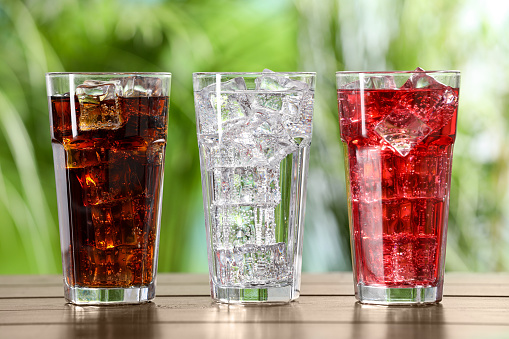 Glasses of different refreshing soda water with ice cubes on wooden table outdoors