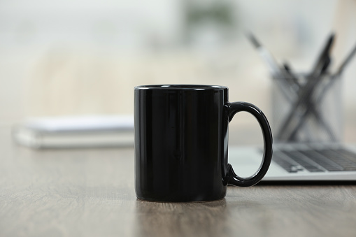 Black ceramic mug and laptop on wooden table indoors. Space for text
