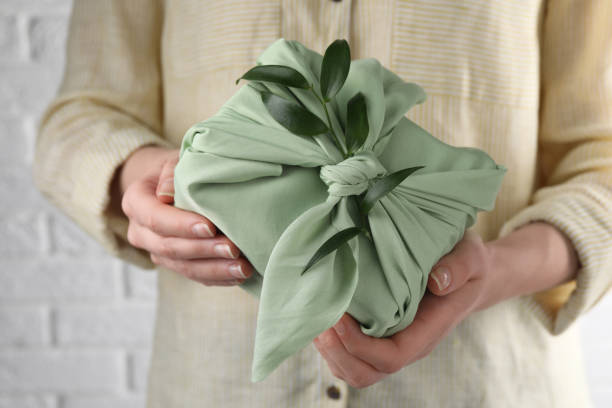 istock Furoshiki technique. Woman holding gift packed in green fabric and decorated with ruscus branch against white wall, closeup 1665971652