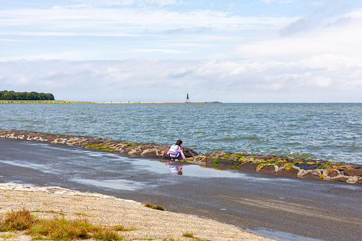 A little girl sitting on the shore of a sea with a lighthouse in the background