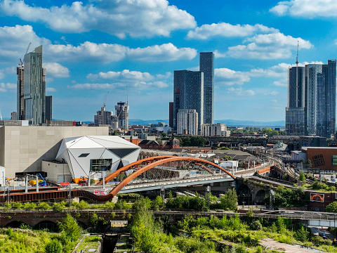 An aerial photograph of Manchester City Centre's modern skyline with skyscrapers both built and some under construction. The Castlefield Curve Railway bridge and rail network can be seen in the foreground. The photograph was produced on a summer day with bright blue skies and fluffy clouds.