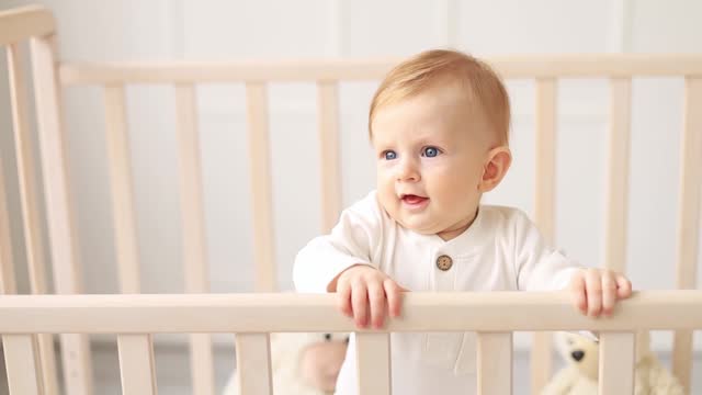 baby boy blond in a white bodysuit stands in a crib holding on to the side of the bed in a bright bedroom and smiles, the morning of the baby