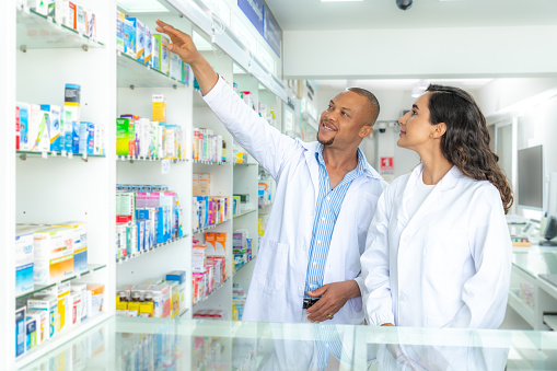 Two multi racial Latin pharmacists, male and female with white lab coat standing next to medicine shelves, finding a medical box for dispensing medicine to patient in a pharmacy.